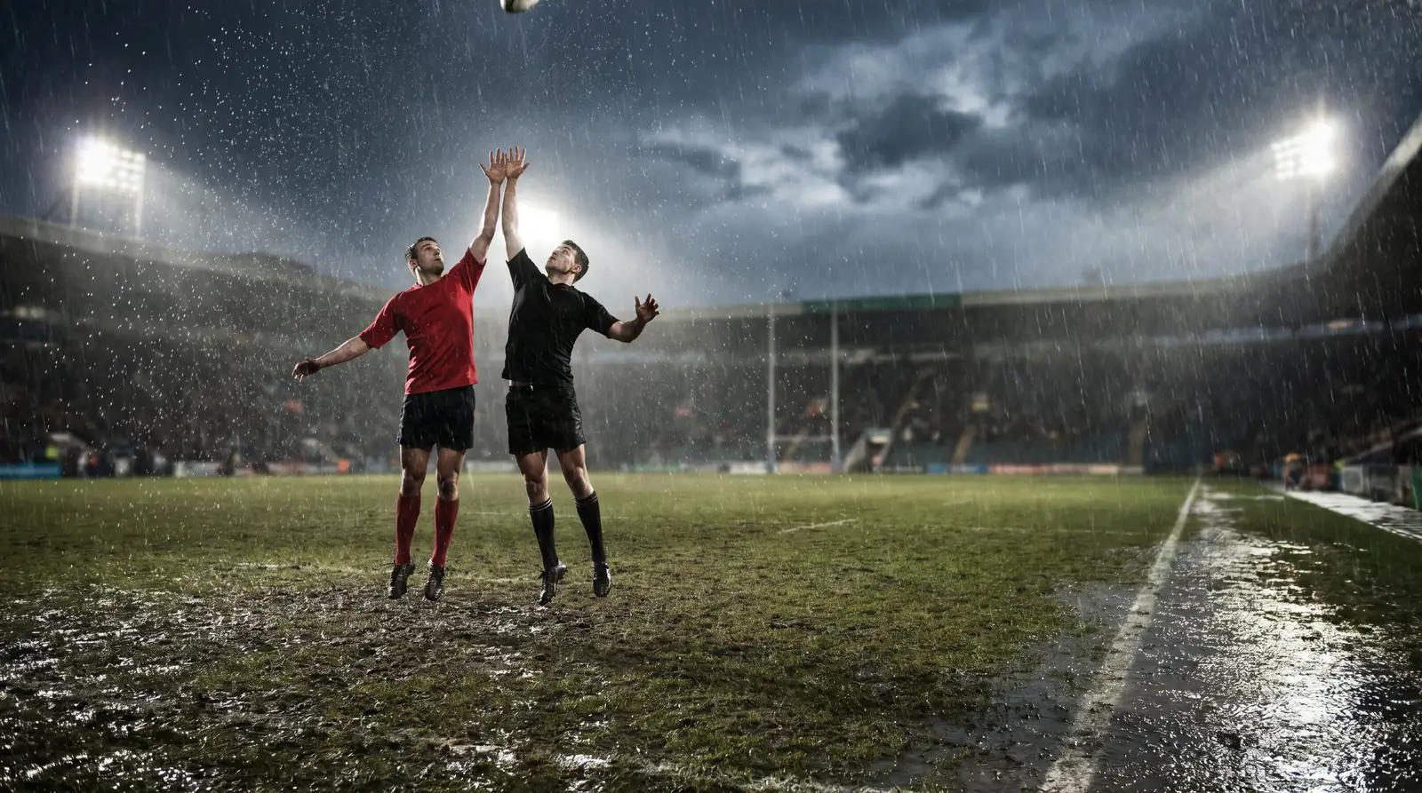 Rugby players competing in heavy rain on a muddy pitch under floodlights