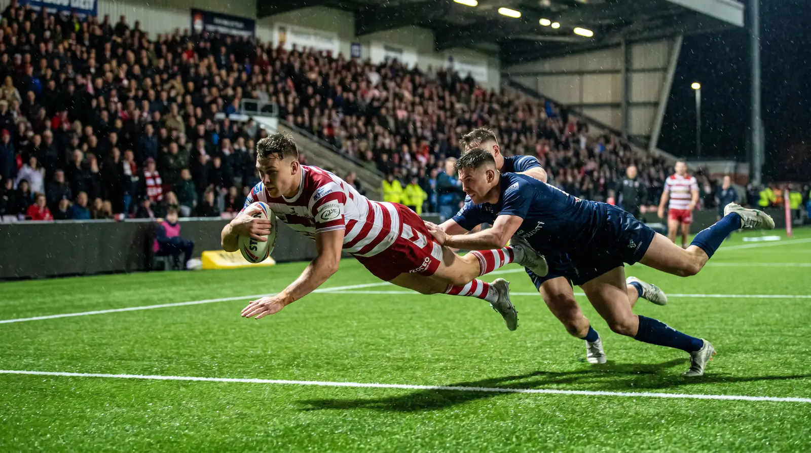 Rugby league player scoring a try with teammates celebrating on a floodlit pitch